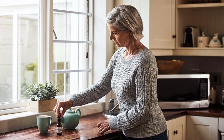 Woman standing in kitchen preparing tea with CBD tincture.