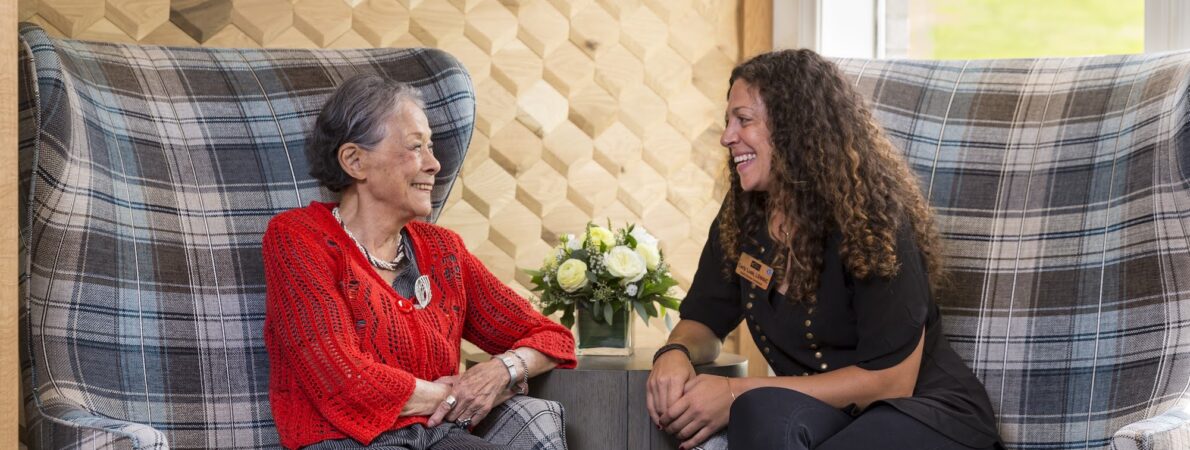 An older lady and a staff member talking and enjoying each others company while sitting in chairs and flowers between them.
