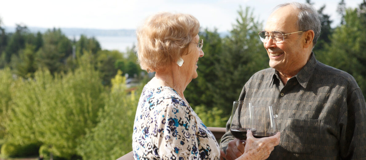 Older couple enjoying wine on a balcony at the Aljoya Mercer Island retirement community in Mercer Island, with a scenic view of the ocean and trees.